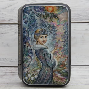 May include: A rectangular metal tin featuring a detailed illustration of a figure wearing a crown and a fur-trimmed coat. The artwork depicts a winter landscape with crystals, snow-covered trees, and white flowers, rendered in shades of blue, purple, and iridescent highlights.