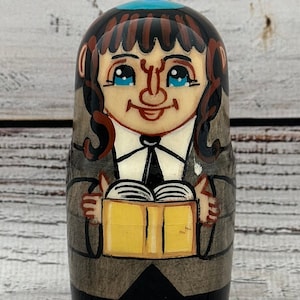 May include: A hand-painted wooden nesting doll featuring a smiling figure with blue eyes, brown hair, and a teal head covering. The doll is holding a yellow book and is dressed in a black suit and gray coat. The background is a light wood grain.