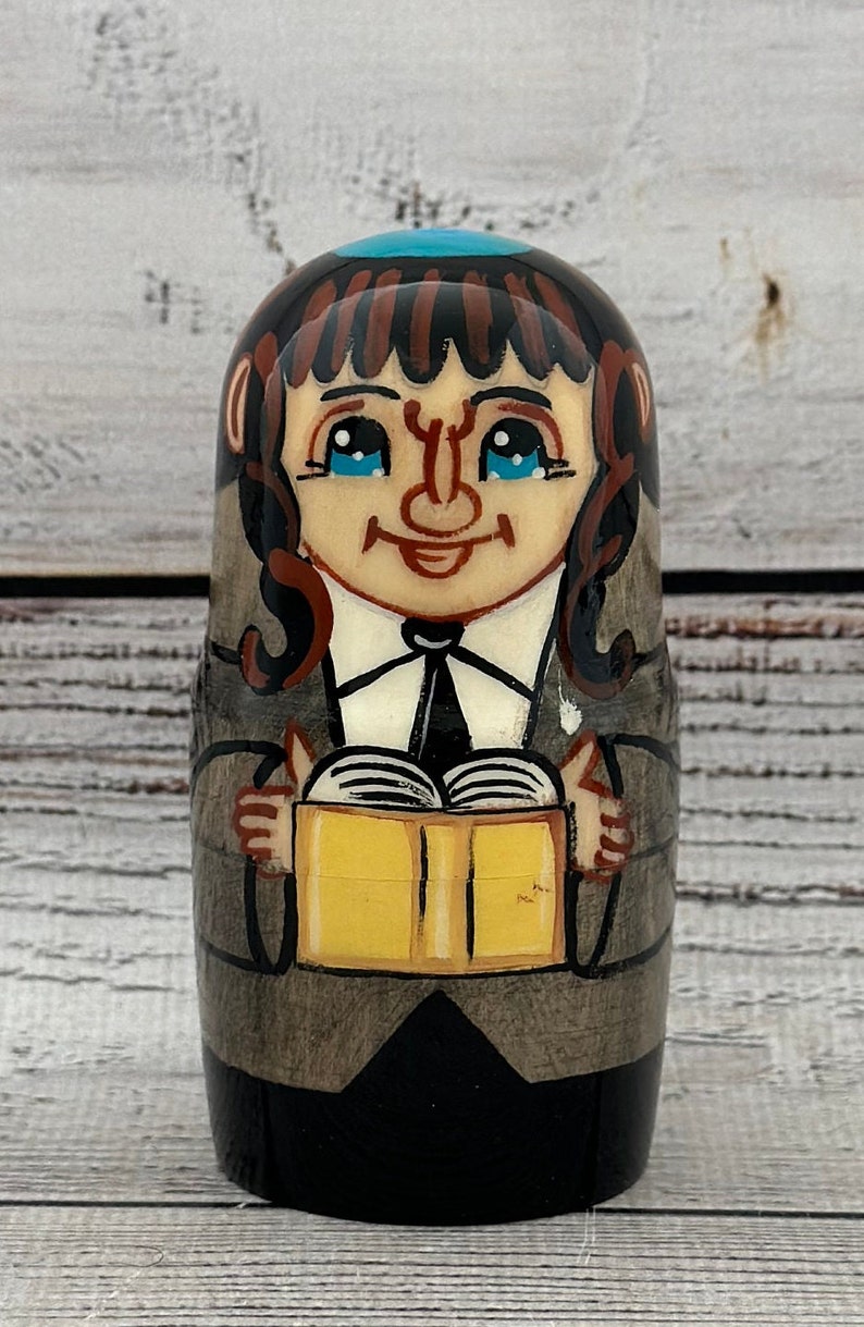 May include: A hand-painted wooden nesting doll featuring a smiling figure with blue eyes, brown hair, and a teal head covering. The doll is holding a yellow book and is dressed in a black suit and gray coat. The background is a light wood grain.