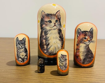 May include: A set of four nesting dolls featuring hand-painted cat portraits. The dolls are cylindrical with rounded tops, painted in shades of beige, brown, and orange. Each doll displays a detailed illustration of a cat within an orange oval frame.