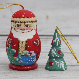 Pode incluir: Dois enfeites de Natal de madeira pintados à mão. Um é uma figura do Papai Noel em vermelho e bege com uma tigela azul de guloseimas. O outro é uma árvore de Natal verde com detalhes em vermelho e branco, ambos com cordões dourados.