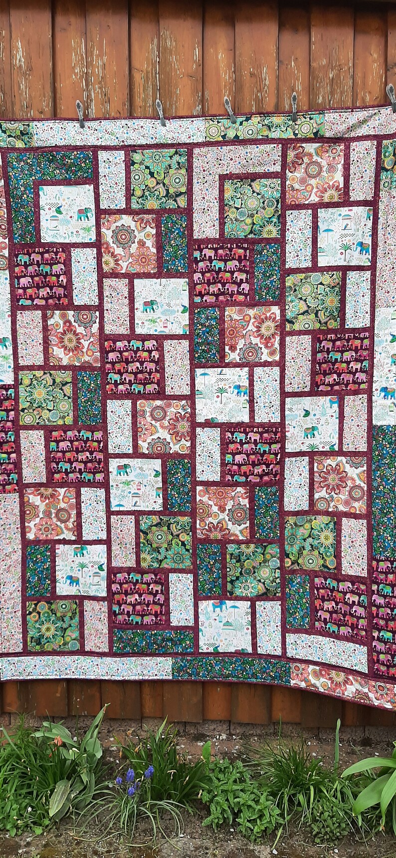 Peut inclure: Une courtepointe patchwork avec un fond bordeaux et diverses cases color&eacute;es repr&eacute;sentant des motifs floraux et d'&eacute;l&eacute;phants.