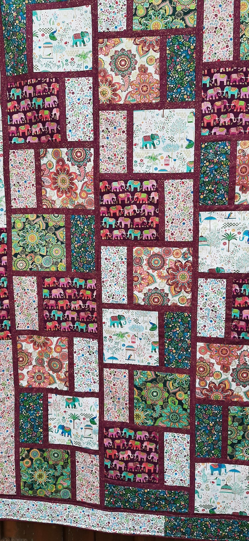 Peut inclure: Une courtepointe compos&eacute;e de diff&eacute;rents carr&eacute;s de tissu pr&eacute;sentant un motif r&eacute;p&eacute;t&eacute; d'&eacute;l&eacute;phants, de paons et de motifs floraux. La courtepointe est r&eacute;alis&eacute;e avec une bordure bordeaux et un fond blanc.