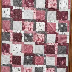 Peut inclure: Une courtepointe en patchwork sur le thème de Noël. La courtepointe est composée de carrés de tissu dans des tons de rouge, gris et blanc. Le tissu présente différents motifs, notamment des flocons de neige, des sapins de Noël et des ornements.