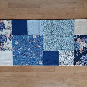 Op de afbeelding: Een blauwe en witte patchwork tafelrunner met een bloemenpatroon. De runner is gemaakt van 12 vierkanten stof, elk met een ander ontwerp. De runner is perfect om elke tafel een vleugje elegantie te geven.