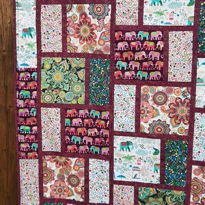 Peut inclure: Une courtepointe en patchwork avec une bordure bordeaux et une vari&eacute;t&eacute; de tissus color&eacute;s repr&eacute;sentant des &eacute;l&eacute;phants, des motifs paisley et des motifs floraux.