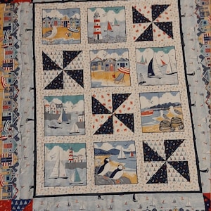 May include: A colourful quilt with a nautical theme. The quilt features a variety of images, including sailboats, lighthouses, and beach houses. The quilt is made up of nine squares, each with a different design. The quilt is bordered by a red, white and blue trim.