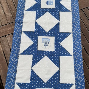 Op de afbeelding: Een blauwe en witte tafelloper met een herhalend sterpatroon. Elke ster heeft een wit midden met een klein geborduurd ontwerp. De ontwerpen omvatten een sneeuwpop, een huis en een boom. De loper heeft een witte achtergrond met blauwe stippen en een blauwe rand met een wit sneeuwvlokkenpatroon.