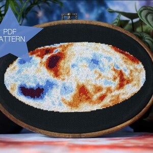 Puede incluir: Un patrón de punto de cruz de una nebulosa colorida en un aro de bordado de madera ovalado. El aro está sobre una mesa con un fondo azul y naranja. Una estrella azul con el texto "PDF PATTERN" está en la esquina superior izquierda de la imagen.
