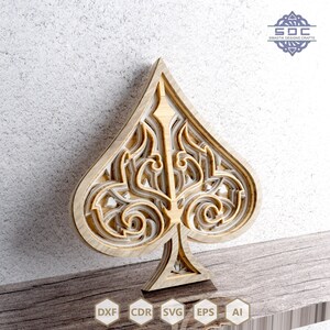 May include: A laser cut wooden design of a spade playing card with intricate details. The design is layered and has a white and brown color scheme.