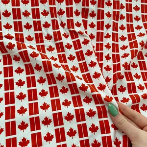 May include: White fabric with a repeating pattern of red maple leaves and red vertical bars. The fabric is slightly creased.