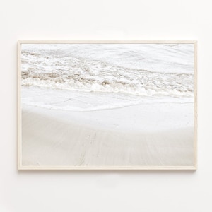 Op de afbeelding: Een ingelijste foto van een strandscène met wit zand en schuimende golven die op het strand breken. De afbeelding is in een lichtbeige kleurenschema.