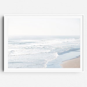May include: A framed photograph of a beach scene with white waves crashing on the shore. The water is a light blue color and the sand is a light brown color. The sky is a light blue color and there are no clouds.