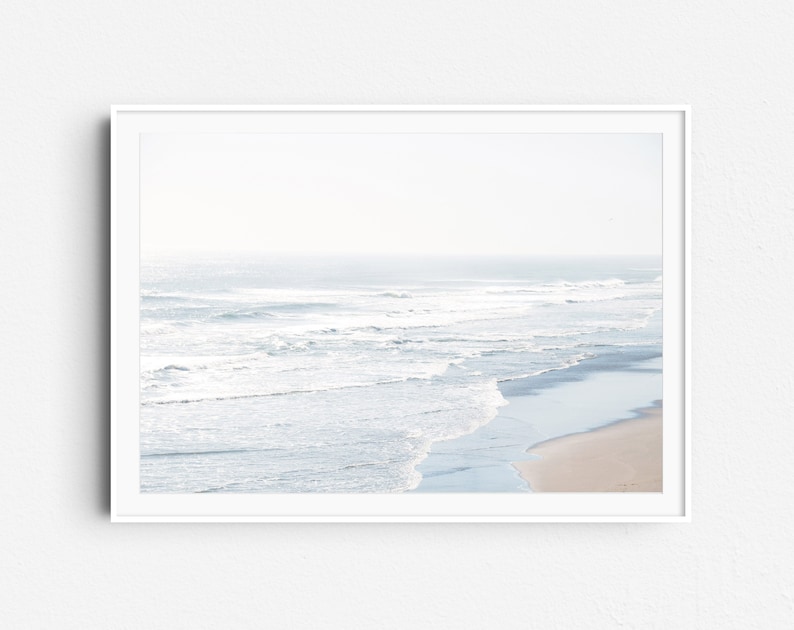 May include: A framed photograph of a beach scene with white waves crashing on the shore. The water is a light blue color and the sand is a light brown color. The sky is a light blue color and there are no clouds.