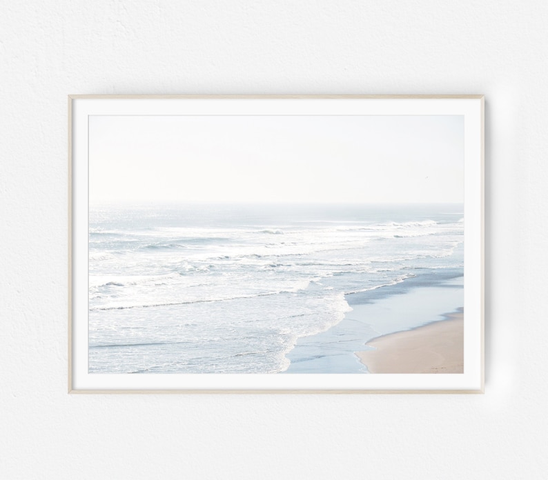 May include: A framed photograph of a beach scene with white waves crashing on the shore. The water is a light blue color and the sand is a light brown color. The sky is a light blue color and there are no clouds visible.