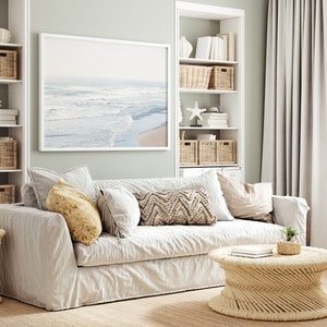 May include: A white linen sofa with pillows in a living room setting. The sofa is in front of a white bookshelf with wicker baskets and a framed photograph of a beach scene. A wicker coffee table sits in front of the sofa.
