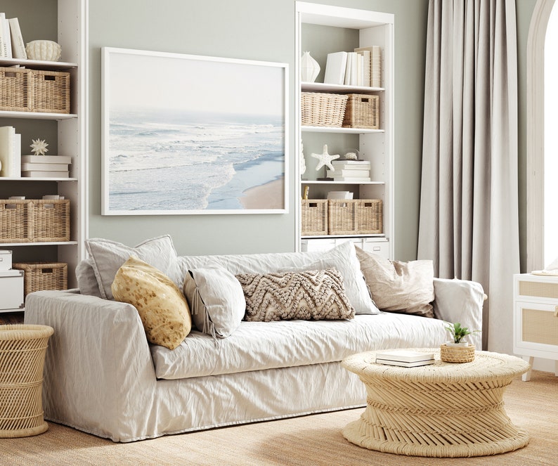 May include: A white linen sofa with pillows in a living room setting. The sofa is in front of a white bookshelf with wicker baskets and a framed photograph of a beach scene. A wicker coffee table sits in front of the sofa.