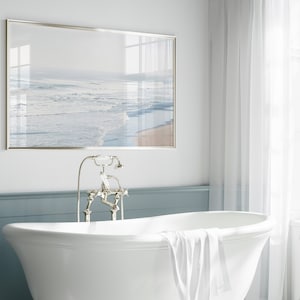 May include: A white bathtub with a white towel draped over the edge. The bathtub is in a bathroom with a framed picture of a beach scene above it and a silver faucet.