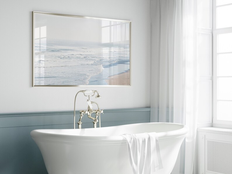 May include: A white bathtub with a white towel draped over the edge. The bathtub is in a bathroom with a framed picture of a beach scene above it and a silver faucet.