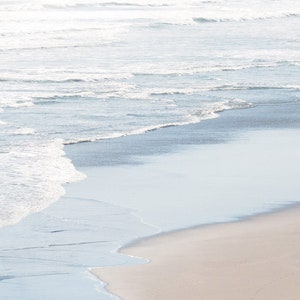 May include: A close-up of a sandy beach with foamy white waves crashing on the shore. The water is a light blue color and the sand is a pale beige.