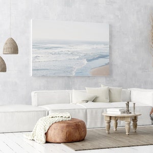 May include: A white sectional sofa with a brown leather ottoman and a white knitted throw blanket. The sofa is in a living room with a white wall, a wooden coffee table, and a canvas print of a beach scene.