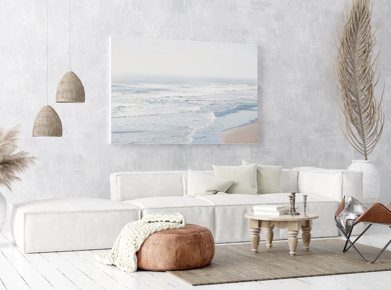 May include: A white sectional sofa with a brown leather ottoman and a white knitted throw blanket. The sofa is in a living room with a white wall, a wooden coffee table, and a canvas print of a beach scene.