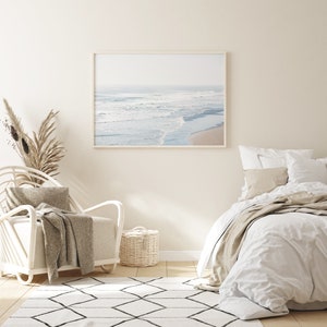 May include: A white bedroom with a white bed, a white wicker chair, a white rug with black diamond patterns, and a framed photograph of a beach scene.
