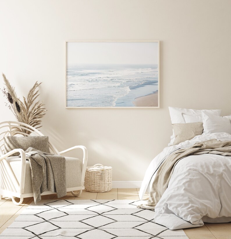 May include: A white bedroom with a white bed, a white wicker chair, a white rug with black diamond patterns, and a framed photograph of a beach scene.