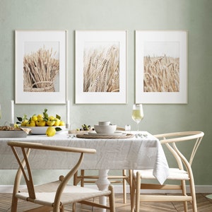 May include: Three framed prints of wheat fields in a light brown color, hanging on a light green wall above a dining table with a white tablecloth, two wooden chairs, and a bowl of lemons.