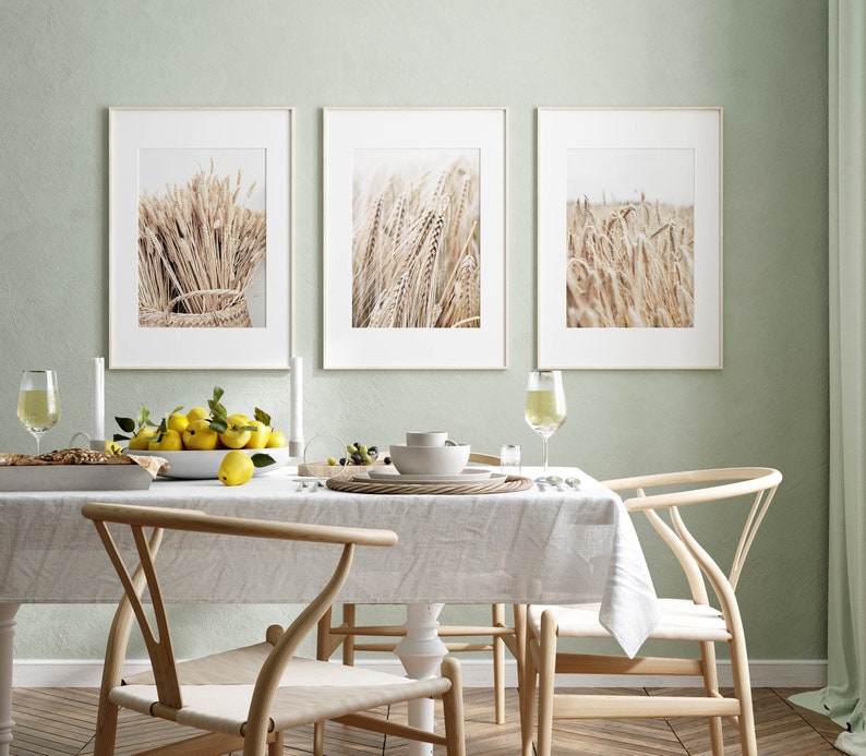 May include: Three framed prints of wheat fields in a light brown color, hanging on a light green wall above a dining table with a white tablecloth, two wooden chairs, and a bowl of lemons.