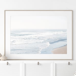 May include: A framed photograph of a beach scene with white waves crashing on the shore. The photo is in a light wood frame and is hanging on a white wall.