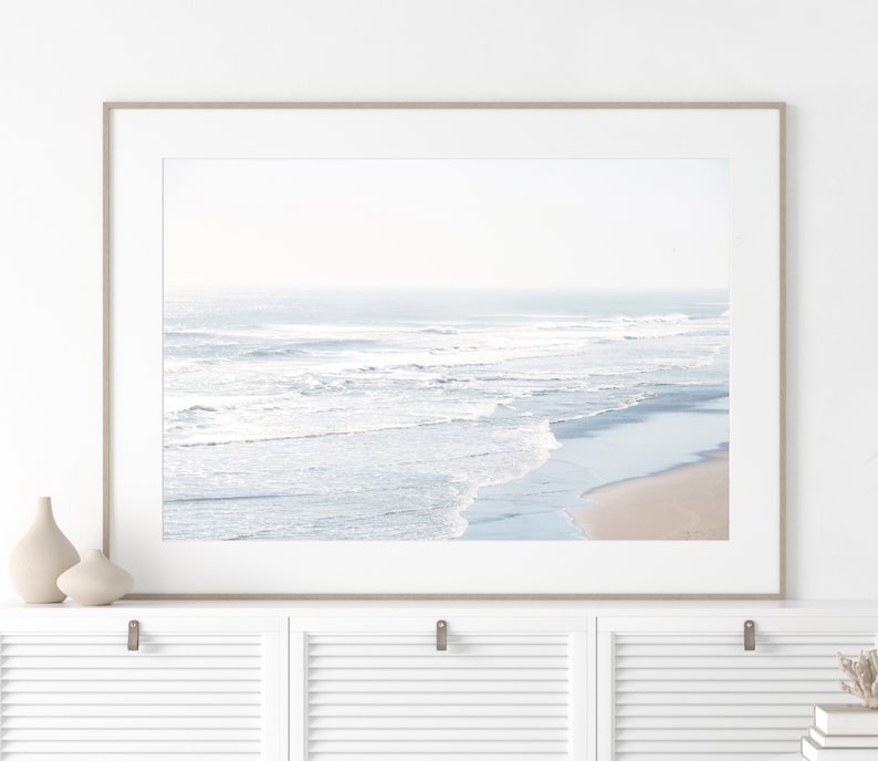 May include: A framed photograph of a beach scene with white waves crashing on the shore. The photo is in a light wood frame and is hanging on a white wall.