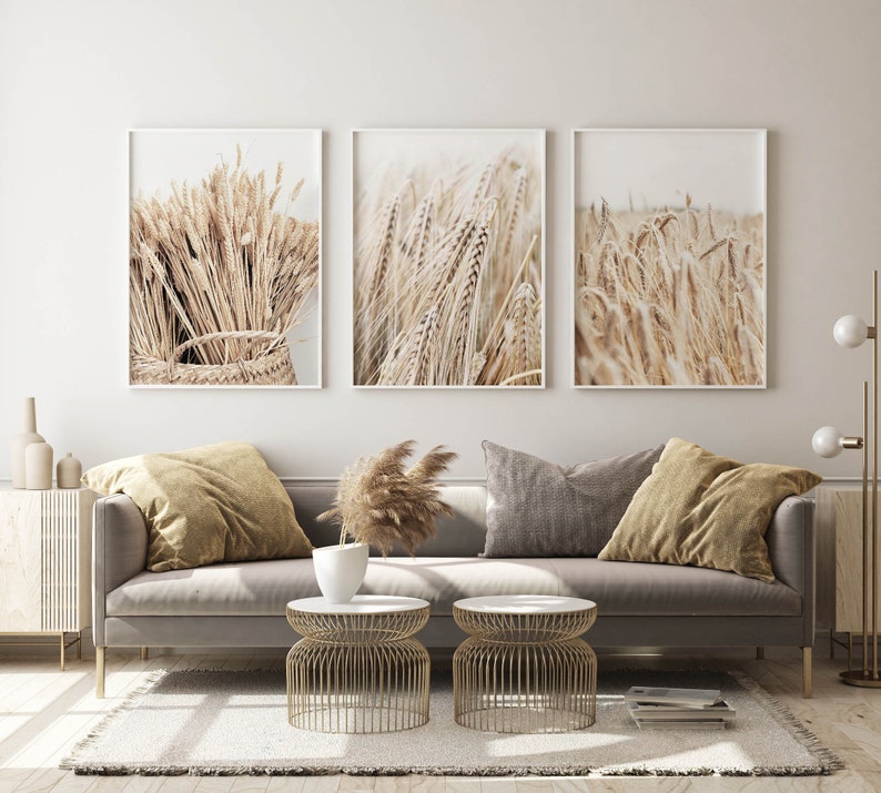 May include: Three framed prints of wheat stalks in a field. The prints are in a living room setting with a gray sofa, two gold side tables, and a vase of dried pampas grass.