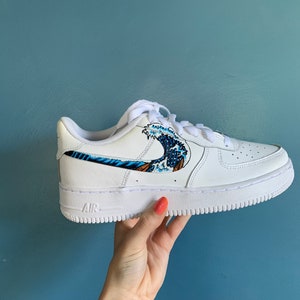 May include: White leather low-top sneakers with a blue and white wave design on the side. The word "AIR" is printed on the sole.