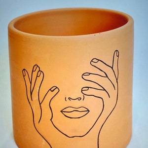 May include: A terracotta planter with a black line drawing of a face with hands covering the eyes. The face has a simple nose and mouth.