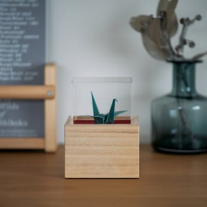 以下が含まれることがあります： 透明なアクリルケースに、ターコイズブルーの折り鶴が展示されています。ケースは木製の自然な仕上げです。折り鶴は、日本の伝統的な紙折り芸術です。