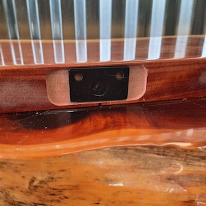 May include: A close-up of a wooden shelf with a black metal bracket attached to the underside. The bracket has two holes and a small metal loop.