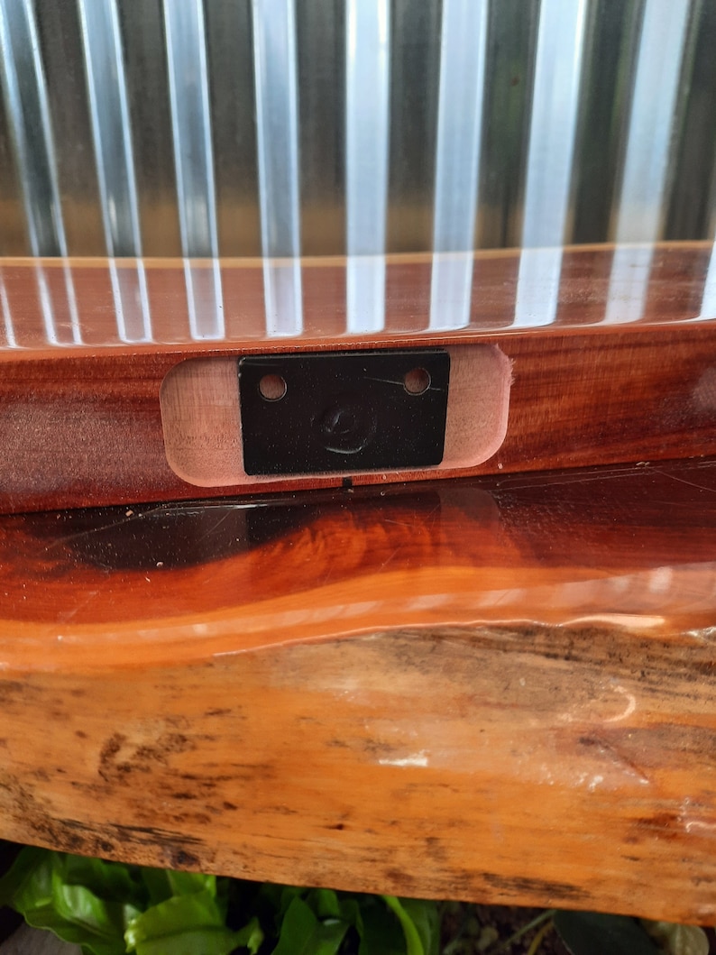 May include: A close-up of a wooden shelf with a black metal bracket attached to the underside. The bracket has two holes and a small metal loop.