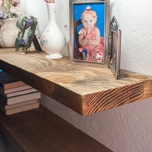 May include: A rustic wooden shelf with a natural wood finish. The shelf is mounted on a white wall and has a few items on it, including a vase of flowers, a picture frame, and a stack of books.