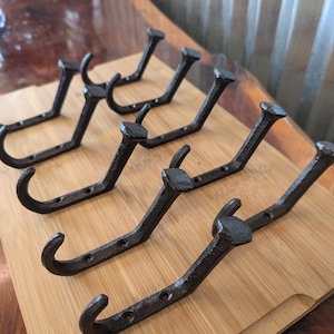 May include: A set of five dark gray metal wall hooks, each shaped like a large railroad spike with a curved end for hanging items. The hooks feature a textured, matte finish and include pre-drilled holes along the base for mounting purposes.
