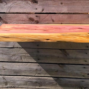 May include: A natural wood shelf with a live edge and a reddish-brown hue. The shelf is mounted on a wooden wall.