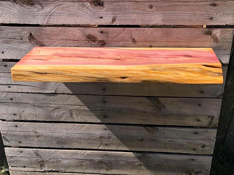 May include: A natural wood shelf with a live edge and a reddish-brown hue. The shelf is mounted on a wooden wall.