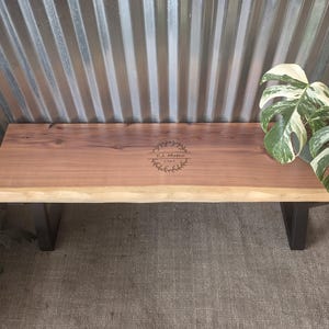 May include: A wooden bench with a live edge and black metal legs. The bench is made of a light brown wood with a natural edge. The text "T.J. Metro 1985" is engraved on the surface. The bench is approximately 60 inches long.