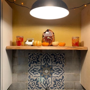 May include: A wooden shelf with a clock made from a tree slice, four small orange pumpkins, and two glass jars with orange and brown contents. The shelf is mounted on a wall with gray tile and a decorative tile with a blue and white pattern.