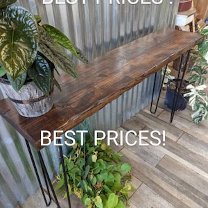 May include: A dark brown wooden console table with a rustic finish and black hairpin legs. The table is sitting in front of a corrugated metal wall with the text "BEST PRICES!" printed on it.