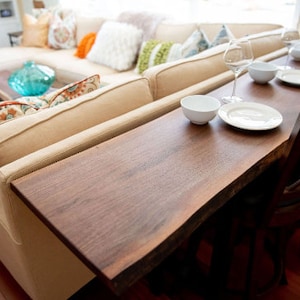 May include: A wooden table with a live edge top and a white chair. The table is set with white plates, bowls, and glasses. The table is in front of a white couch with pillows.