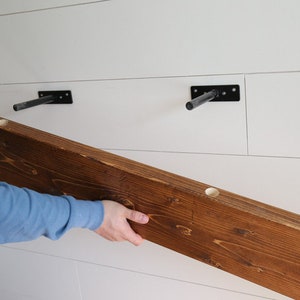 May include: A person is installing a wooden shelf with two black metal brackets. The shelf is stained a dark brown color and has three pre-drilled holes.