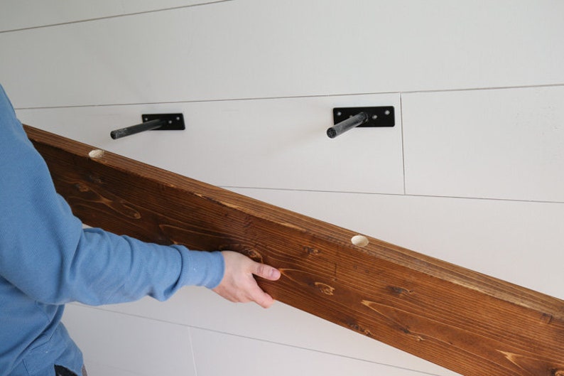 May include: A person is installing a wooden shelf with two black metal brackets. The shelf is stained a dark brown color and has three pre-drilled holes.