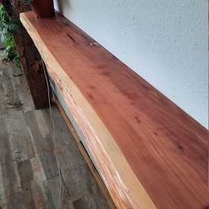 May include: A long, wooden shelf with a natural edge and a reddish-brown finish. The shelf is mounted to a white wall and has a light brown wooden support underneath.