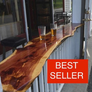 May include: A wooden bar top with a live edge, mounted on a white railing. Two glasses of beer are on the bar top. The bar top is made of natural wood with a reddish brown color. The text "BEST SELLER" is in red letters on a white background.